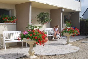 A building exterior with white benches, potted plants with red flowers, and a small tree. - F1-JARDINET privé CLOS - PARKING privé /MER à 300 Mètres Plages du Débarquement  (Asnelles)
