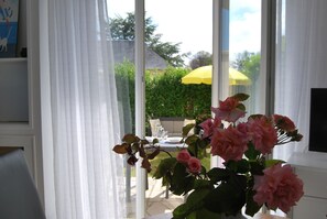 A room with a large window, a table with chairs, and a vase with pink flowers. - F1-JARDINET privé CLOS - PARKING privé /MER à 300 Mètres Plages du Débarquement  (Asnelles)