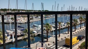 A balcony with a table featuring a glass of wine and a placemat with a heart design. In the background, there is a marina with boats and a clear blue sky. - Gruissan (Aude). Quiet apartment with marina view and parking space (Gruissan)