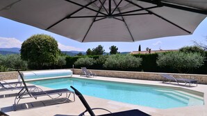 Ein Pool mit Liegestühlen und Sonnenschirm, umgeben von Steinmauern und Grünpflanzen, mit Blick auf Berge im Hintergrund.