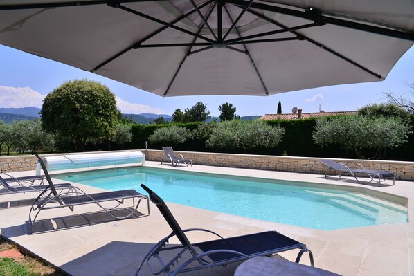 A swimming pool with lounge chairs and an umbrella, surrounded by stone walls and greenery, with a view of mountains in the background. - Cottage facing the luberon with swimming pool (Apt)
