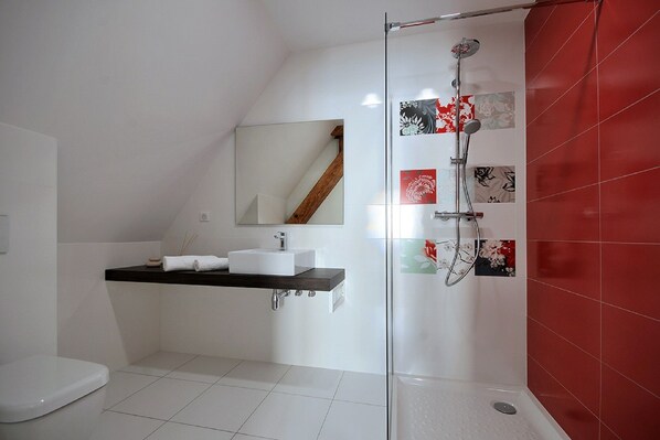 Une salle de bain moderne avec une douche à parois de verre, des carreaux blancs, une étagère en bois avec un lavabo blanc et un mur de douche recouvert de carreaux rouges.