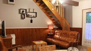 Ein Wohnzimmer mit einem braunen Ledersofa, einem Holztisch und einem Fernseher auf einer Holzkommode. Im Hintergrund ist eine Treppe mit Holzbalkonen zu sehen.