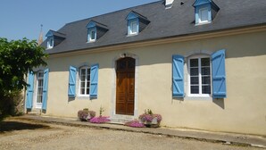 Un bâtiment à deux étages doté d’une façade symétrique, avec une porte en bois au centre, des volets bleus et une cheminée.