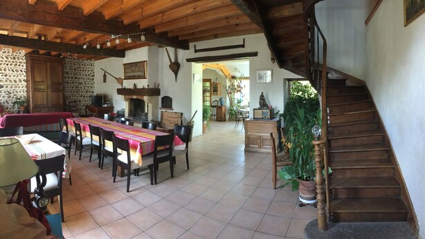 Un coin repas rustique avec une longue table, des chaises et une cheminée. Un escalier en bois mène à un niveau supérieur.