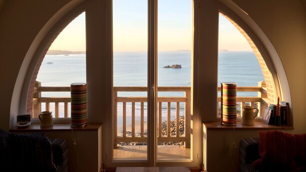 A room with a large arched window overlooking a beach, a wooden railing, and a table with a striped vase and a kettle. - Superb comfortable apartment with sea view (Perros-Guirec)