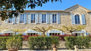 A stone building with multiple windows and shutters, surrounded by well-maintained hedges and potted plants.