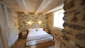 A bedroom with a stone wall, wooden ceiling beams, a bed with white bedding, two bedside tables, and two wall-mounted lights.
