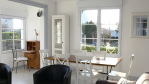 Habitación luminosa con paredes blancas, una mesa de comedor de madera y sillas blancas. Se observa una silla negra en primer plano, un aparador de madera y una ventana con vistas a zonas verdes.