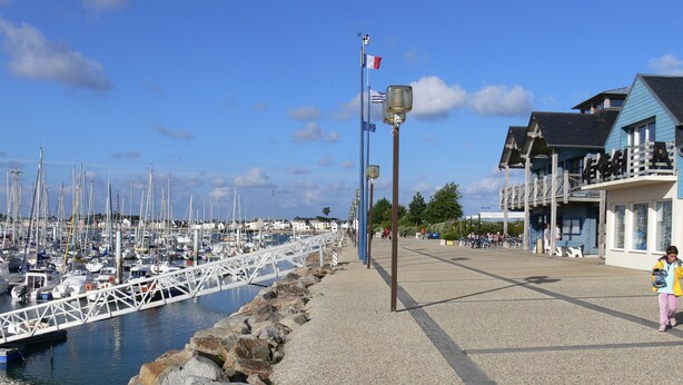 Un puerto deportivo con barcos amarrados, un paseo peatonal y un edificio de fachada azul.