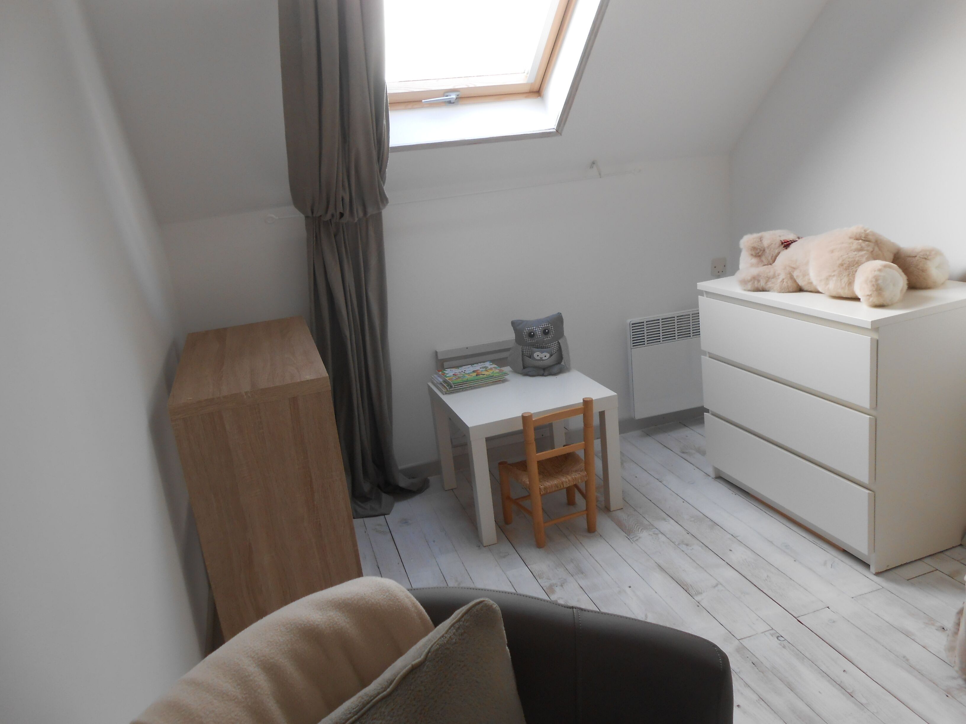 A small, bright room with a skylight, a wooden desk, a chair, a white dresser, and a teddy bear on top.