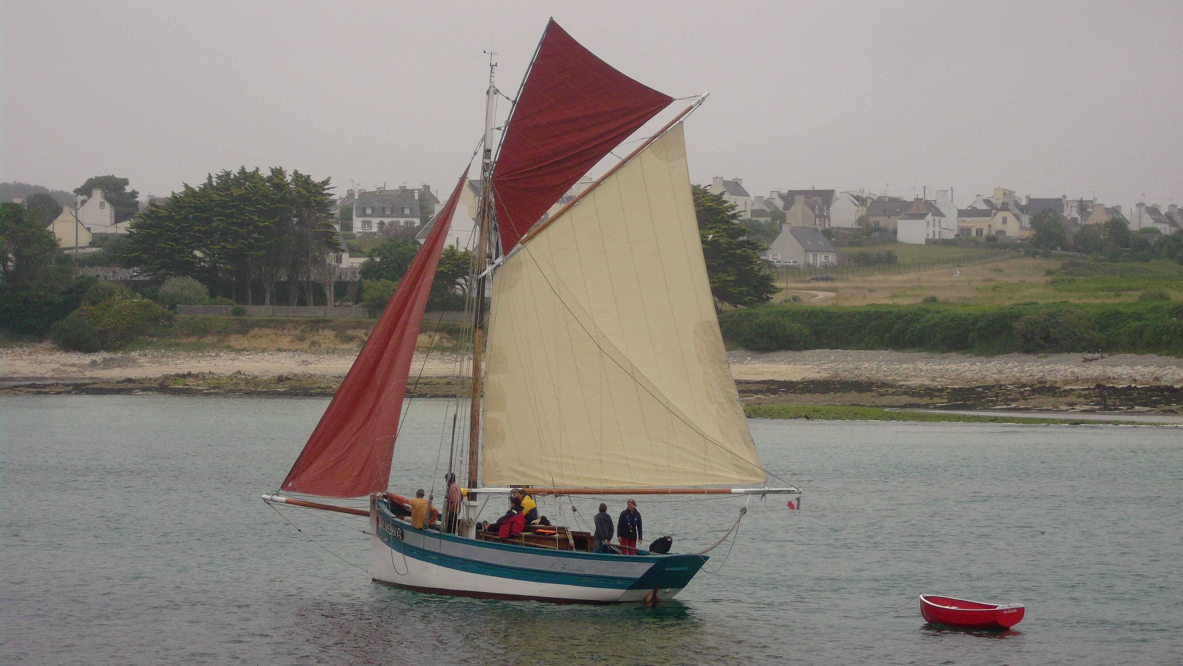 A sailboat with a red and white sail on calm water, surrounded by a residential area with houses and greenery.