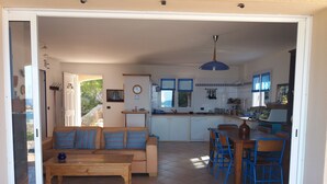 A spacious living area with a sofa, wooden coffee table, and a dining table with chairs. A kitchen with a sink, stove, and cabinets is visible in the background. - villa marina , climatised,wi-fi, private garden and parking. (Sartène)