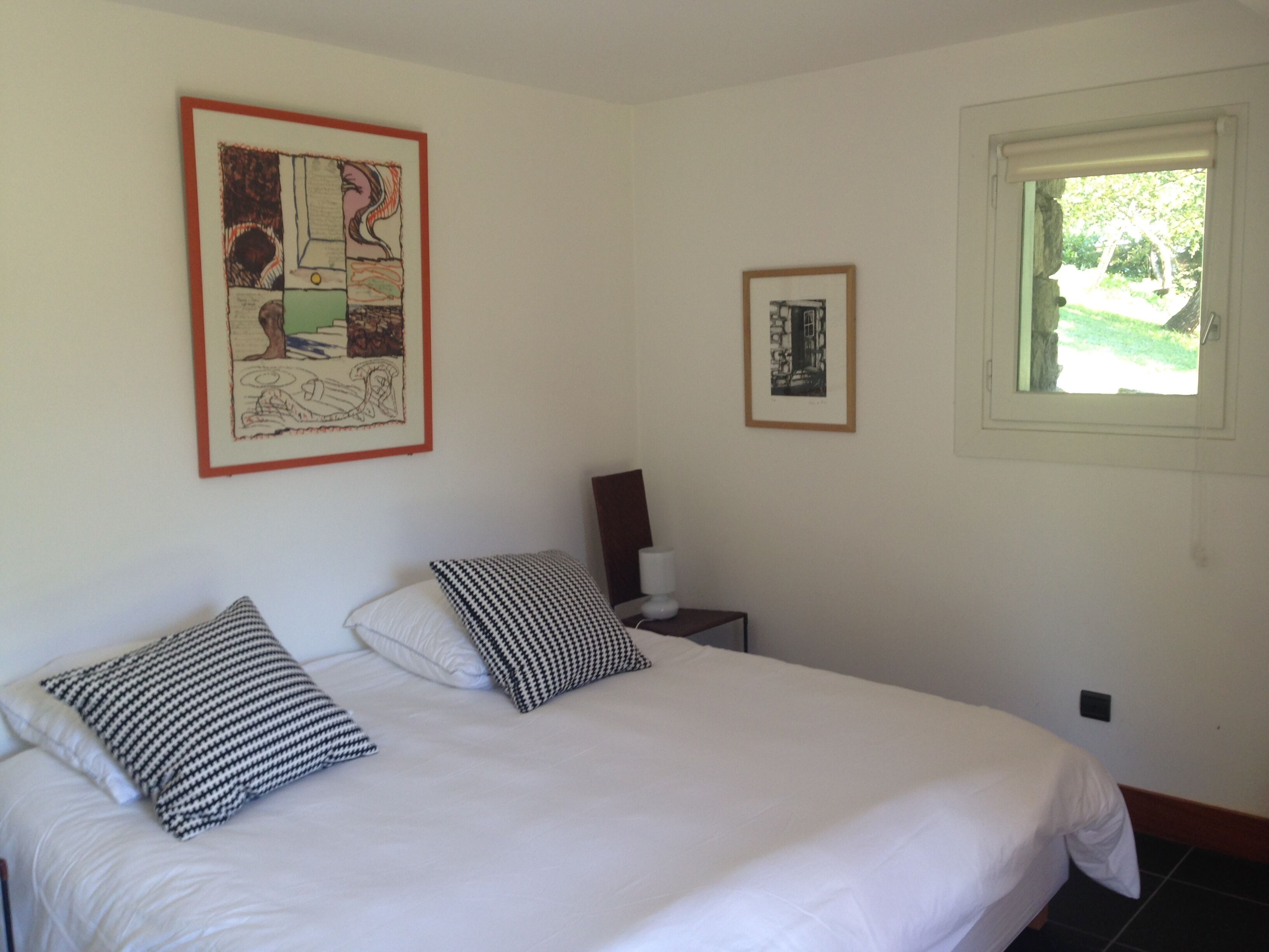 Een slaapkamer met een groot bed, twee kussens, een nachtkastje met een lamp en twee ingelijste kunstwerken aan de muur.