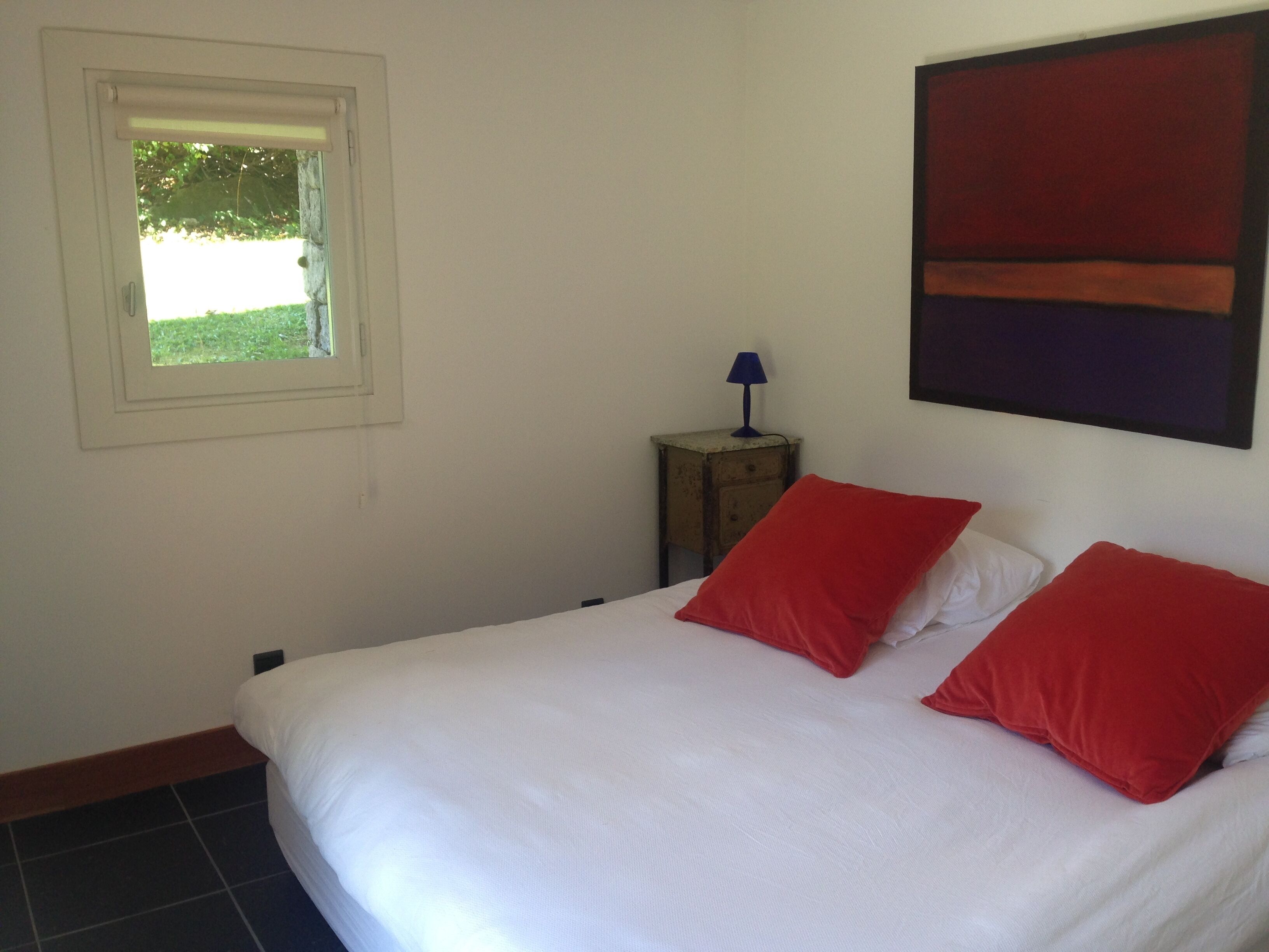 Une chambre à coucher avec un lit, deux oreillers rouges, une table de chevet avec une lampe et une fenêtre donnant sur un paysage verdoyant.