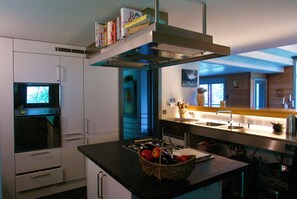 A modern kitchen with a built-in oven, a microwave, and a range hood. There is a basket of fruit on the counter and a sink with a faucet.