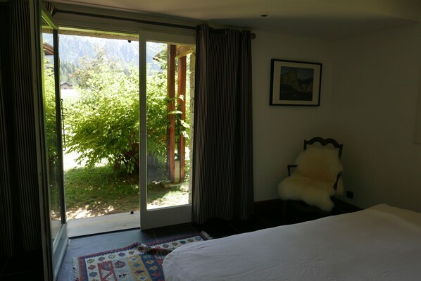 A bedroom with a bed, a chair, a painting on the wall, and a view of greenery through the door.
