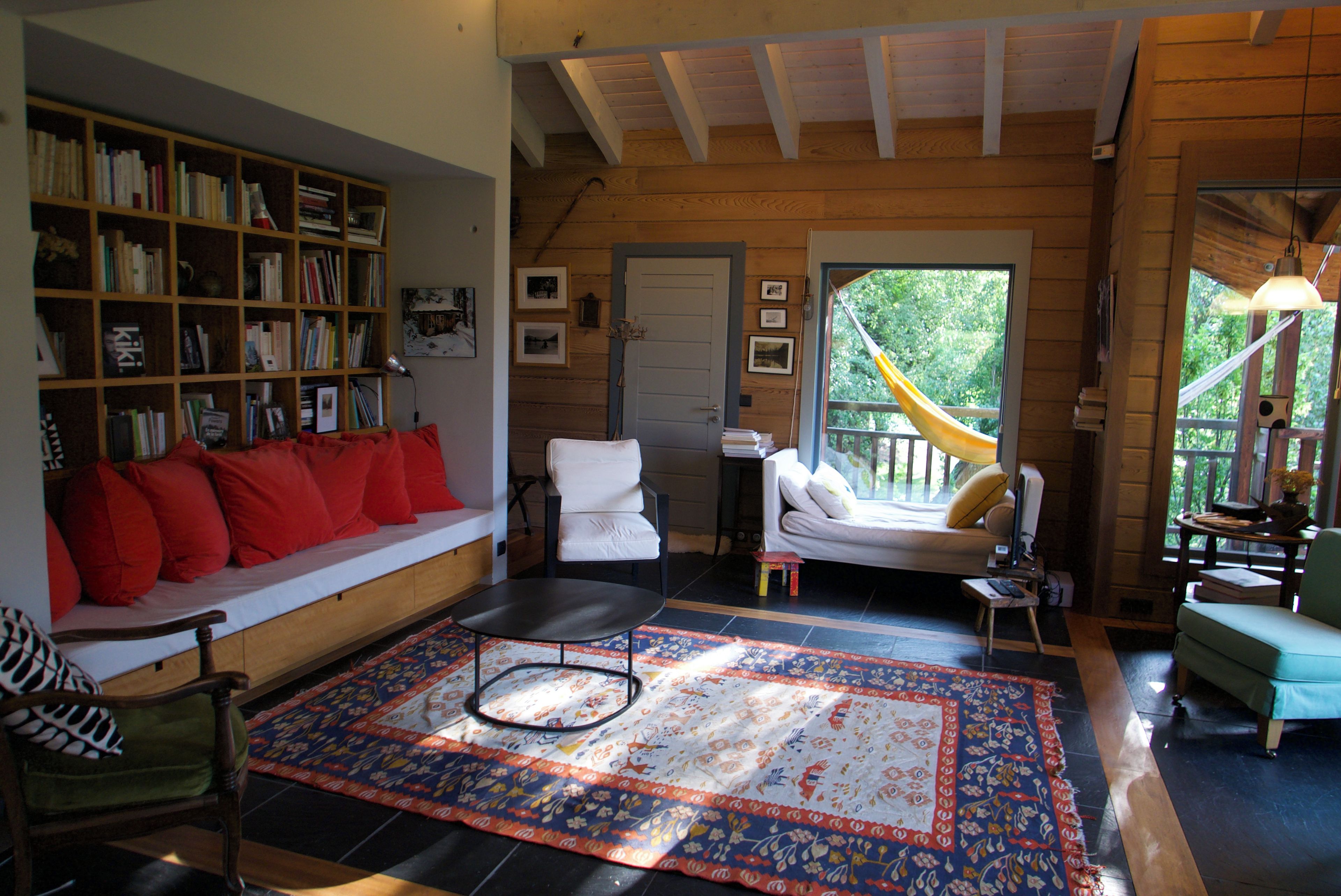 Un salon chaleureux avec une bibliothèque, un canapé orné de coussins rouges, un tapis à motifs et une vue sur l’extérieur par une porte.