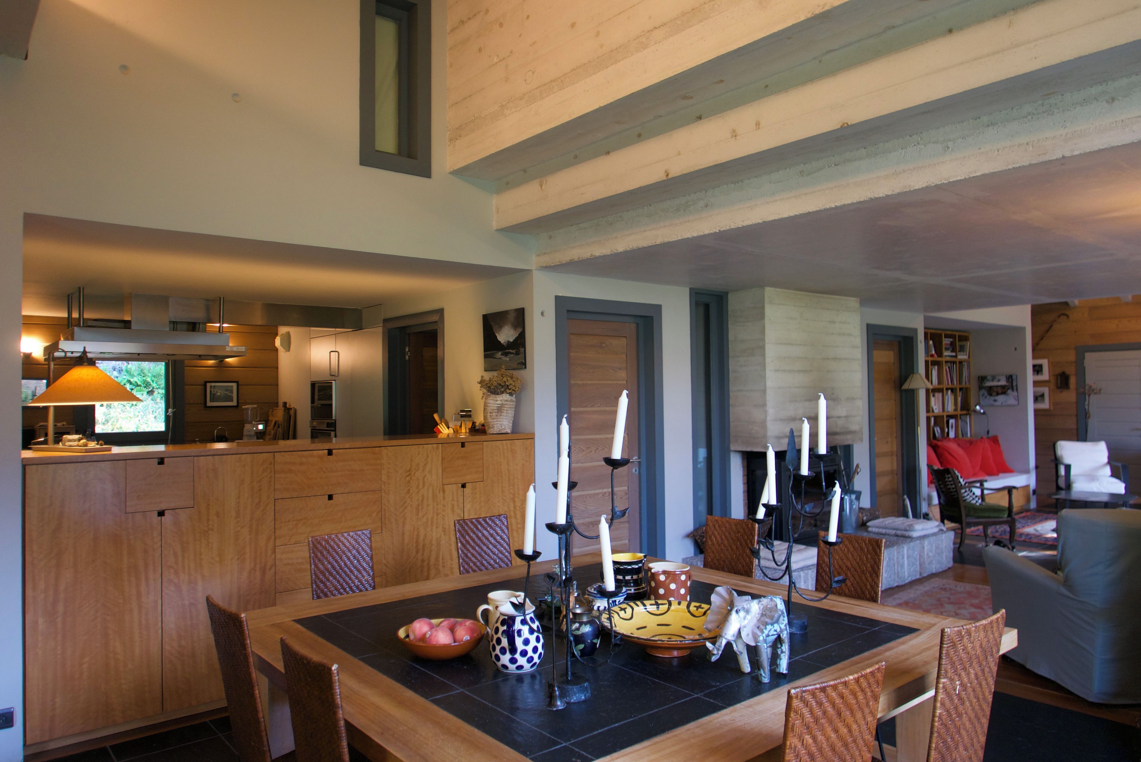Un vaste espace de vie comprend une table à manger en bois, entourée de chaises en osier. On aperçoit, au fond, une cuisine équipée d’appareils électroménagers modernes.