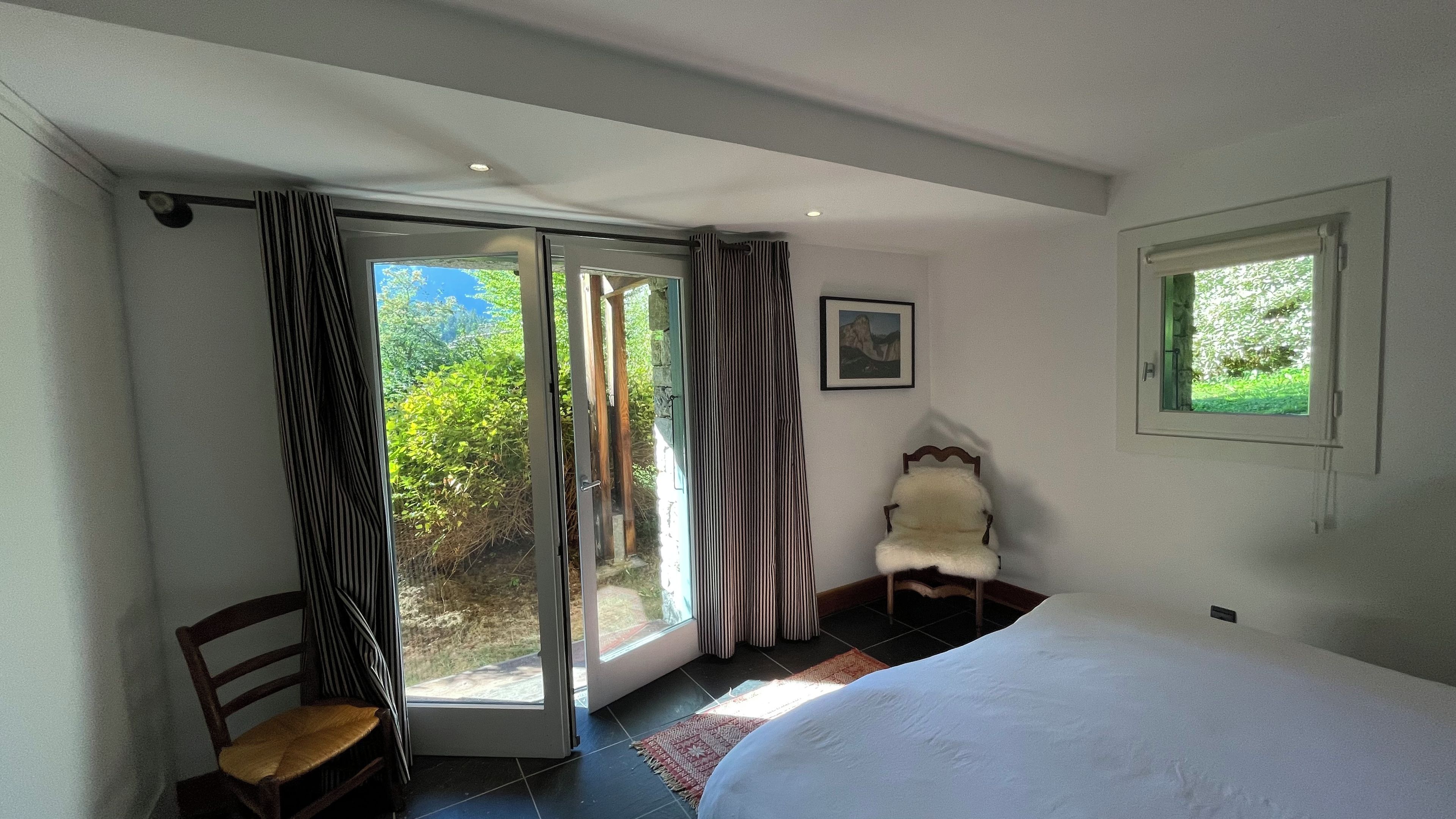 Een slaapkamer met een bed, een stoel, een raam en een deur die uitkomt in een tuin.