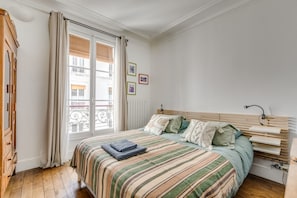 A bedroom with a bed, a wooden headboard, a bedside table, a lamp, and a window with curtains.