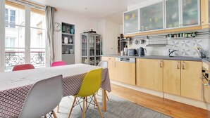 A modern kitchen with a dining area, featuring a table with chairs, a cabinet with glass doors, and a view of the outside balcony. - Cosy and spacious 145m² family apartment (Paris)