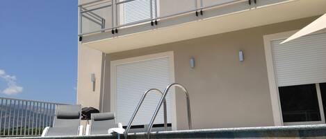 A swimming pool area with a metal ladder, lounge chairs, and a building with a balcony.