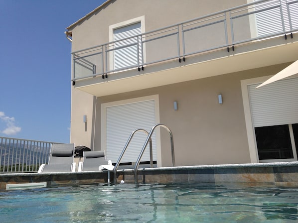 A swimming pool area with a metal ladder, lounge chairs, and a building with a balcony.