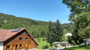 A wooden chalet with a sloped roof and a balcony, surrounded by a lush green landscape and a mountain backdrop.