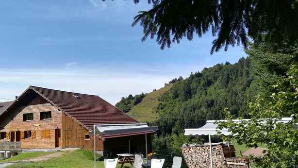A wooden building with a sloped roof, a covered outdoor seating area with chairs and tables, and a stone wall.