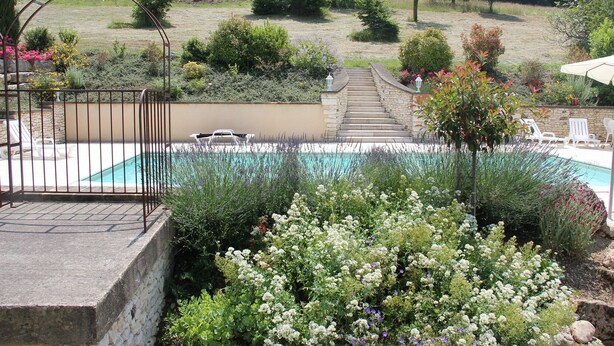 Un espace piscine agrémenté d’un escalier en pierre menant à une terrasse, le tout entouré d’une végétation luxuriante et de plantes en fleurs.