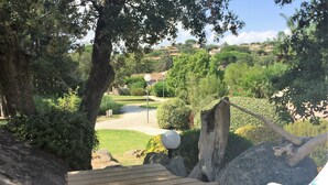 A wooden deck with a white chair and a blue cushion, overlooking a landscaped garden with a pathway and trees. - 100 metres from Santa Guilia beach, comfortable, garden furniture, barbecue (Porto-Vecchio)