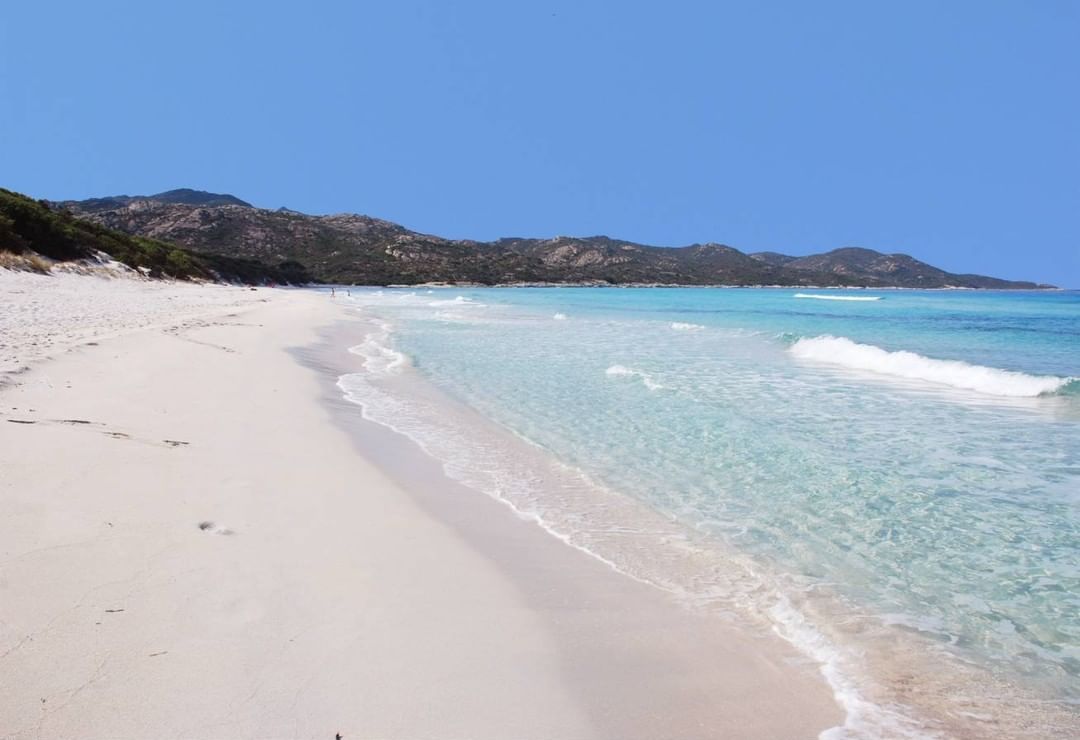 Ein Strand mit klarem türkisblauem Wasser, Sandstrand und in der Ferne Bergen.
