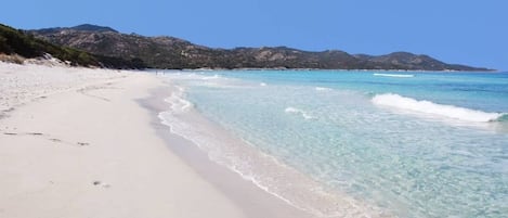 Ein Strand mit klarem türkisblauem Wasser, Sandstrand und in der Ferne Bergen.