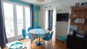 A dining area with a round table, blue chairs, and a striped tablecloth. There is a bookshelf with books and a TV on the wall. - Apartment 40 m2, very comfortable, south facing, Le Tréport, full center and beach (Le Tréport)