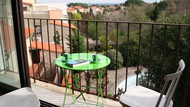 Un balcon avec une table verte et deux chaises blanches, donnant sur un quartier résidentiel avec des maisons aux toits rouges et de la verdure.
