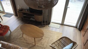 Un salon comprenant un canapé, une table basse ronde, un fauteuil en osier, un téléviseur posé sur un meuble en bois, et une grande fenêtre donnant sur une terrasse en bois.