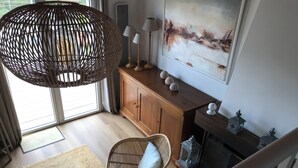 Un salon avec un fauteuil en osier, un meuble en bois, une cheminée et un grand tableau accroché au mur.