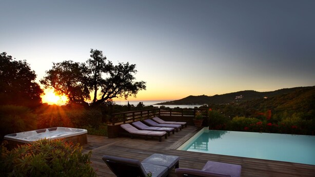 Terraza de madera con sillones de descanso y jacuzzi con vista a una piscina y al atardecer.