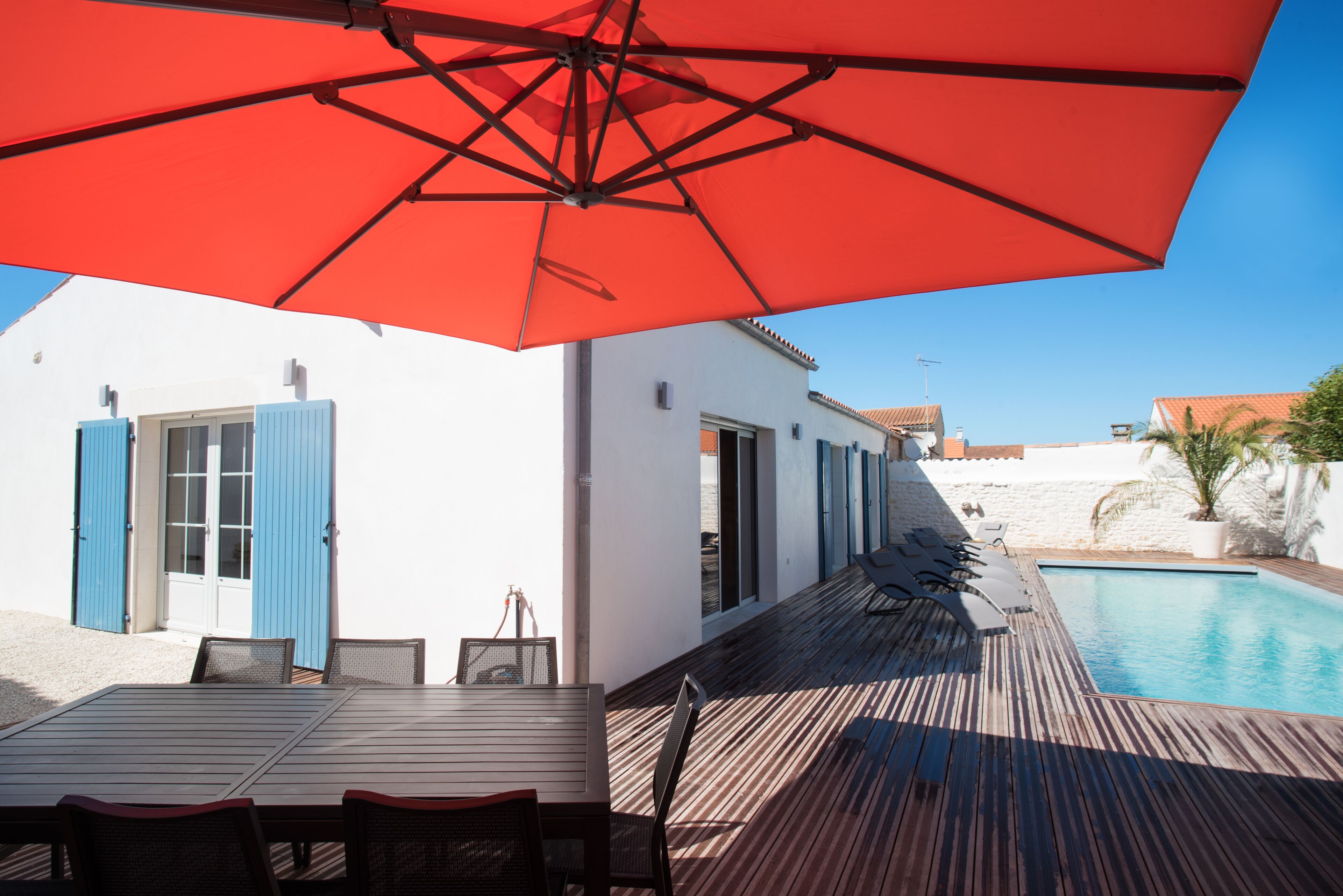 Um deck de madeira com piscina, uma mesa com cadeiras e um guarda-sol vermelho.