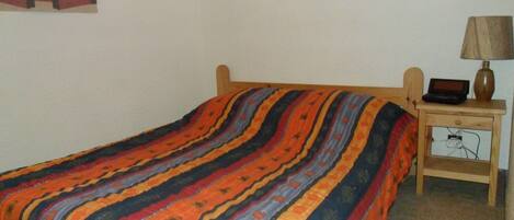 A single bed with a colorful striped bedspread, a wooden headboard, and a bedside table with a lamp and a small electronic device.
