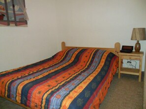 A single bed with a colorful striped bedspread, a wooden headboard, and a bedside table with a lamp and a small electronic device. - Holiday Apartment (Orcières)