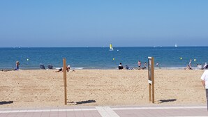 Ein Strand mit Menschen, die entspannen und sonnenbaden, ein klarer, blauer Himmel und ein ruhiges Meer mit einigen Booten in der Ferne.
