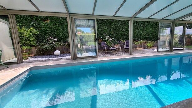 Ein hallenbad mit großen Glaswänden, einer Holzdielen-Terrasse und Blick auf einen Garten mit Pflanzen und einem Zaun.