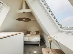 A bedroom with a sloped ceiling, a bed, a bedside table with a vase, and a view through a window. - Le Manoir du Four à Chaux (Paimpol)