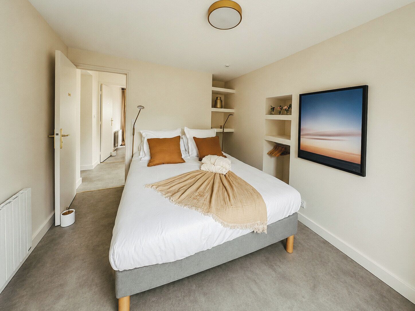 A bedroom with a bed, a bedside table, a lamp, a shelf with decorative items, and a painting on the wall.