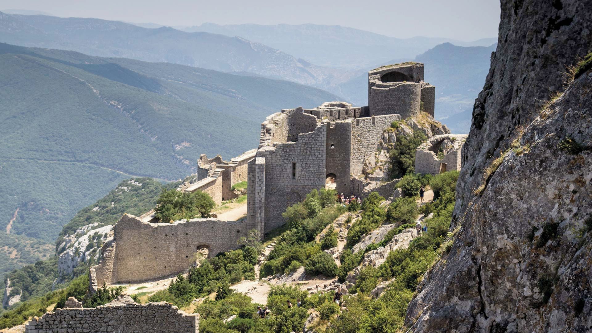 An ancient stone fortress with ruins, surrounded by lush greenery and mountains in the background.