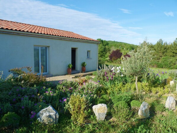 A house with a tiled roof, a front door, and a small window. There are potted plants on the porch and a garden with various plants and flowers in front of the house. - Charming villa in the heart of the hills of the Aude Pyrenees foothills (Donazac)