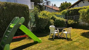 Un espace extérieur verdoyant avec un toboggan vert, deux chaises en plastique blanches et une petite table ronde.