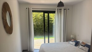 Ein Schlafzimmer mit einem großen Bett, einer hängenden Leuchte, einem Spiegel und Blick auf einen Garten durch eine Schiebeglastür.